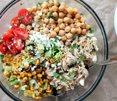 a glass bowl of mixed greens topped with chopped chicken, chickpeas, corn, tomato, avocado, and cheese