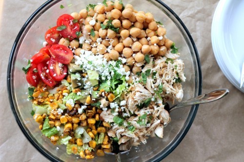a glass bowl of mixed greens topped with chopped chicken, chickpeas, corn, tomato, avocado, and cheese