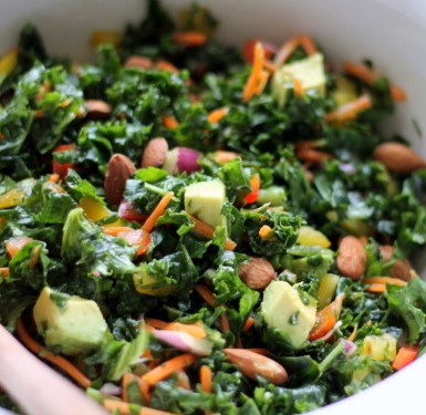 A white bowl filled with kale salad with tons of other veggies like onion, carrots. brussels sprouts, and almonds