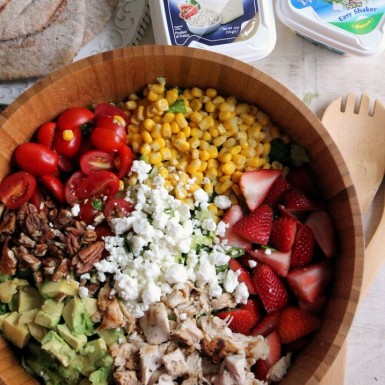 a large wooden bowl filled with salad covered in sections of strawberries, avocado, feta, corn, and nuts