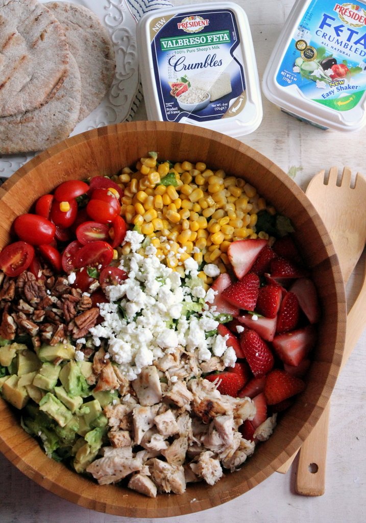 a large wooden bowl filled with salad covered in sections of strawberries, avocado, feta, corn, and nuts