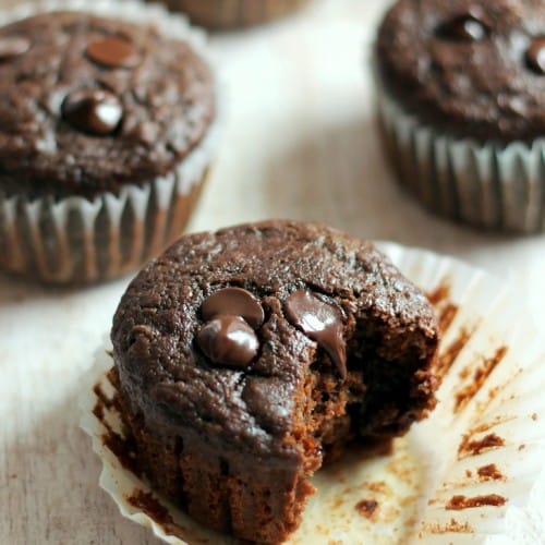 Double Chocolate Zucchini Muffins on a white table. One of the four muffins has a bite taken out of it.