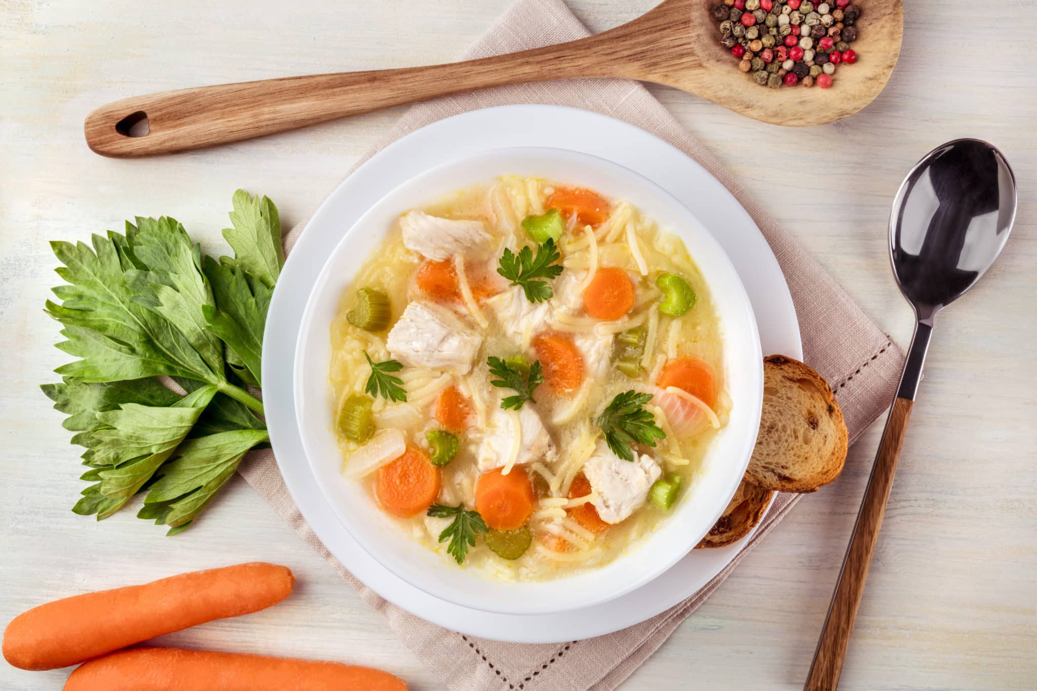 Bowl of chicken soup with vegetables on table with large carrots and wooden spoon