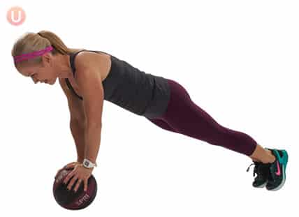 Chris Freytag demonstrating a medicine ball plank with a medicine ball wearing a black top and workout pants.