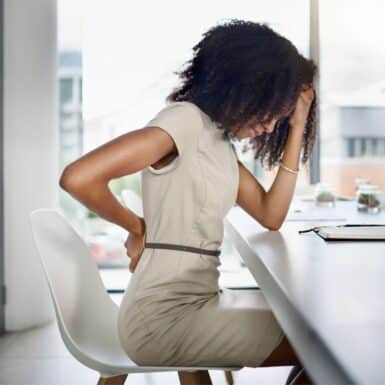 A woman sitting at a desk with her head in one hand and her other clutching her lower back.