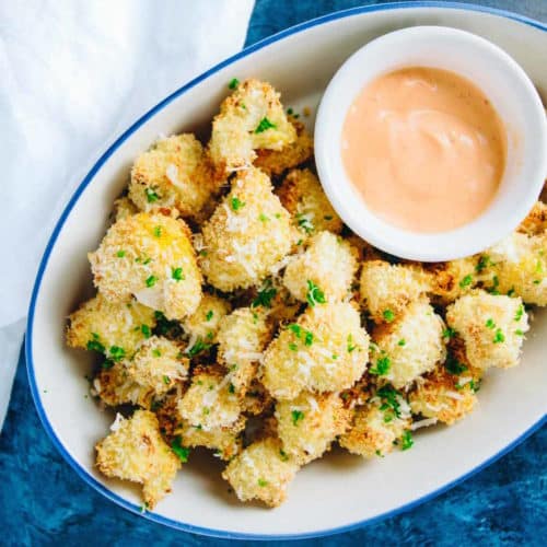 Crispy cauliflower bites in a bowl with a side of sriracha sauce on blue background