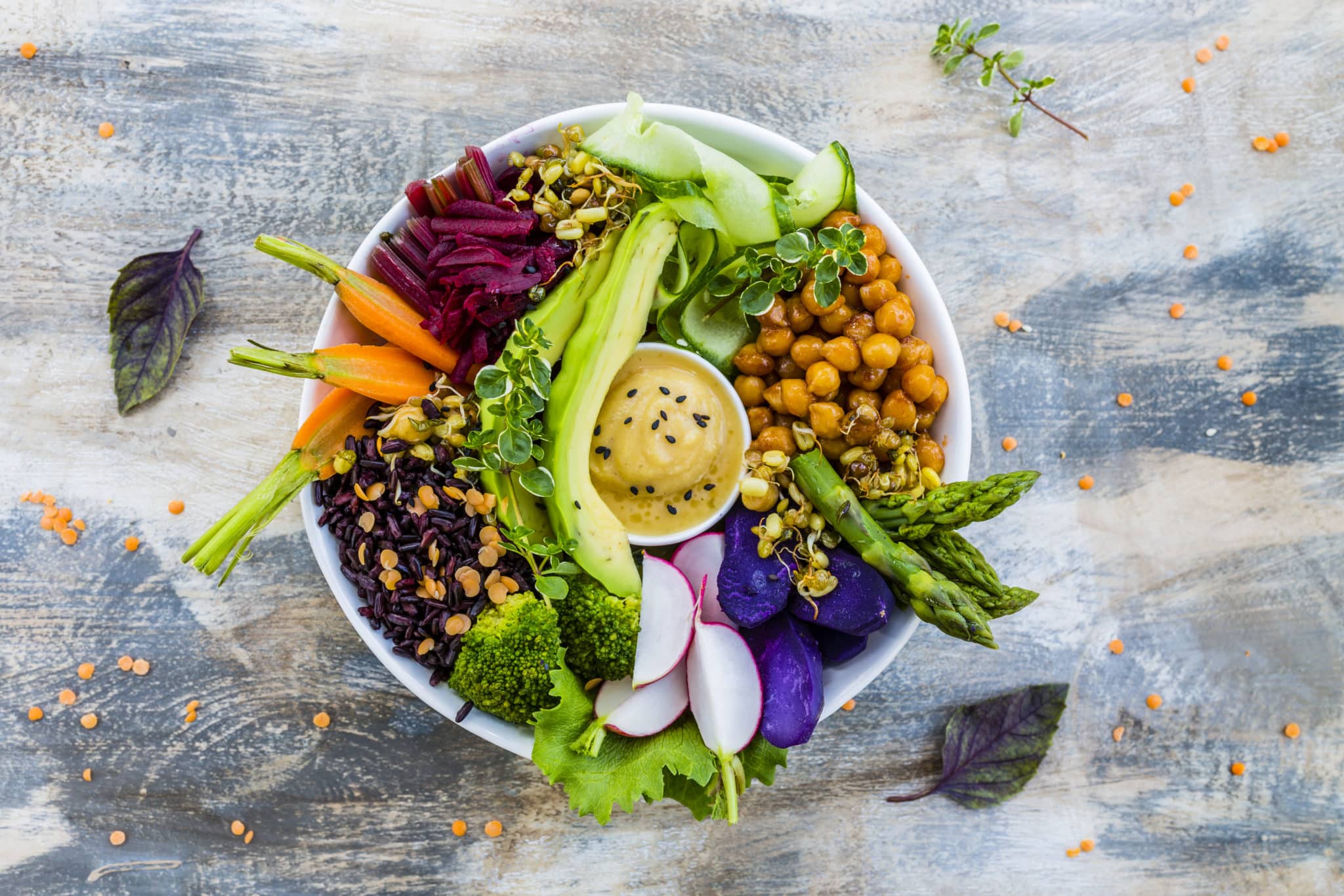 Vegetarian buddha bowl on wood table with carrots, black rice, lentils, hummus, avocado and more