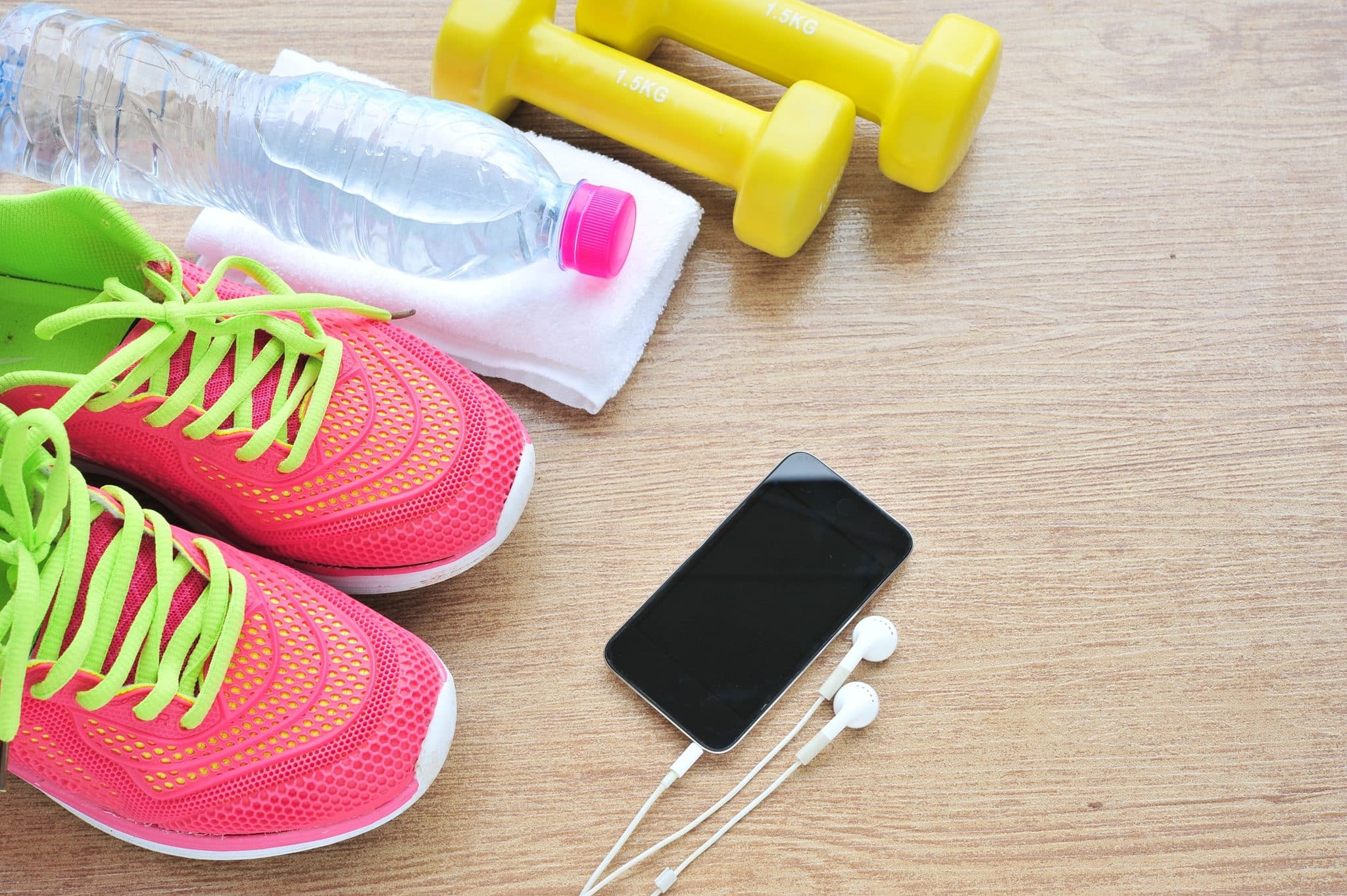 A group of items on the floor, including a pair of pink and green running shoes, a water bottle, two yellow dumbbells, and a smartphone with attached headphones.