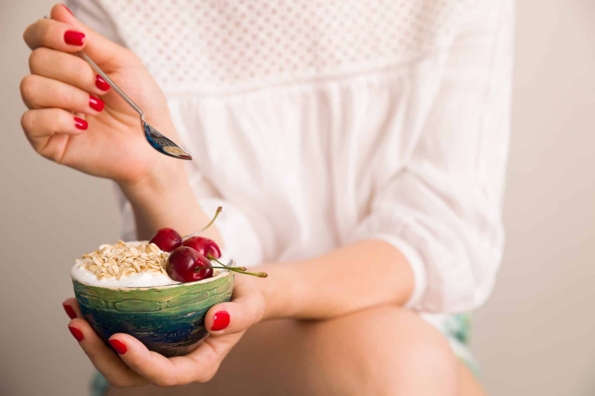 Closeup of woman's hands holding a cup with organic yogurt with oats and cherries in green bowl.