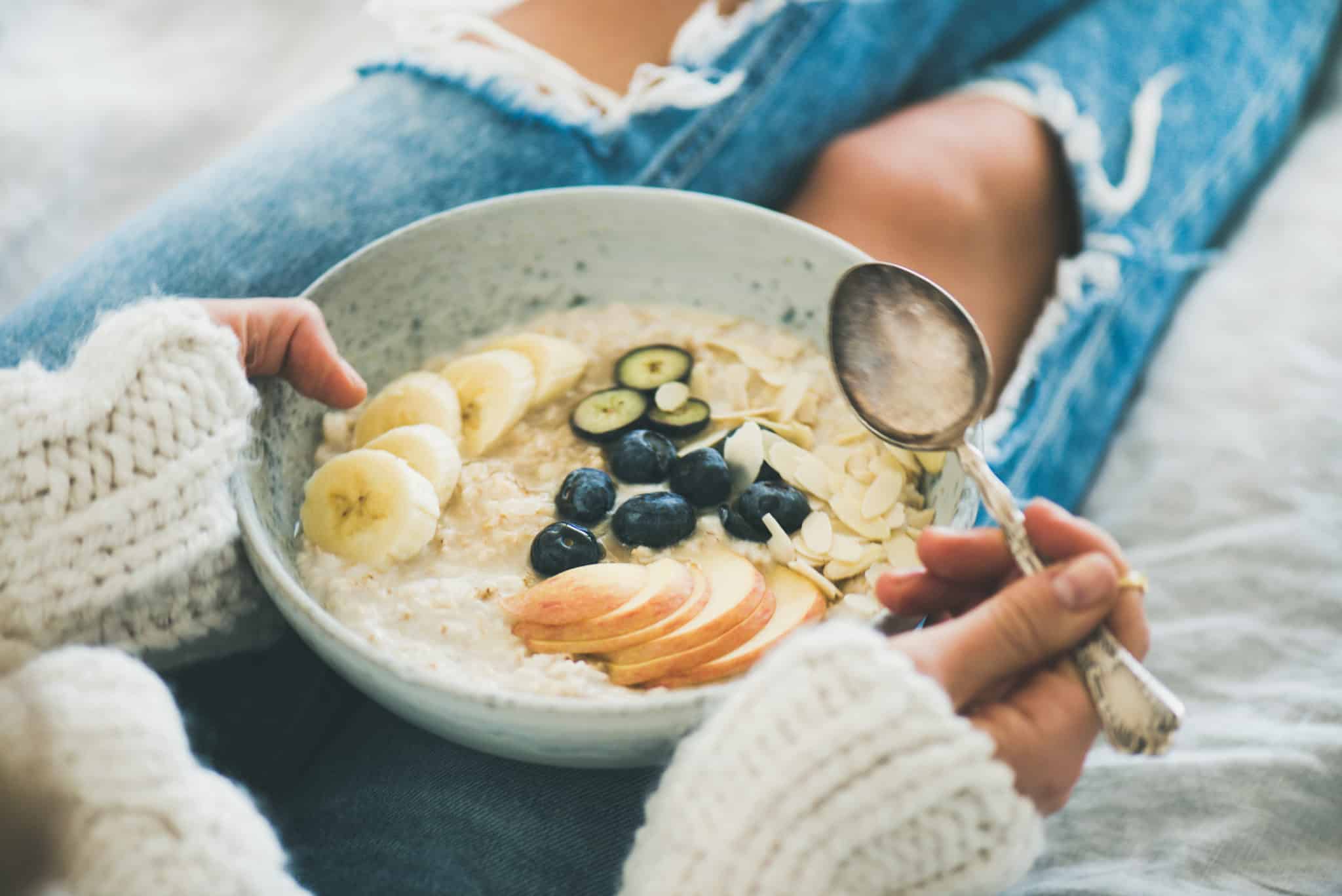 Woman in woolen sweater and shabby jeans eating vegan almond milk oatmeal in bowl with berries, fruit and almonds.