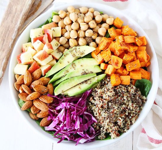 Beautiful big salad in white bowl containting chickpeas, almonds, sweet potatoes, avocado, purple cabbage, apples quinoa and chia seed dressing