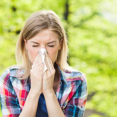 A woman sneezing into a tissue in front of trees.