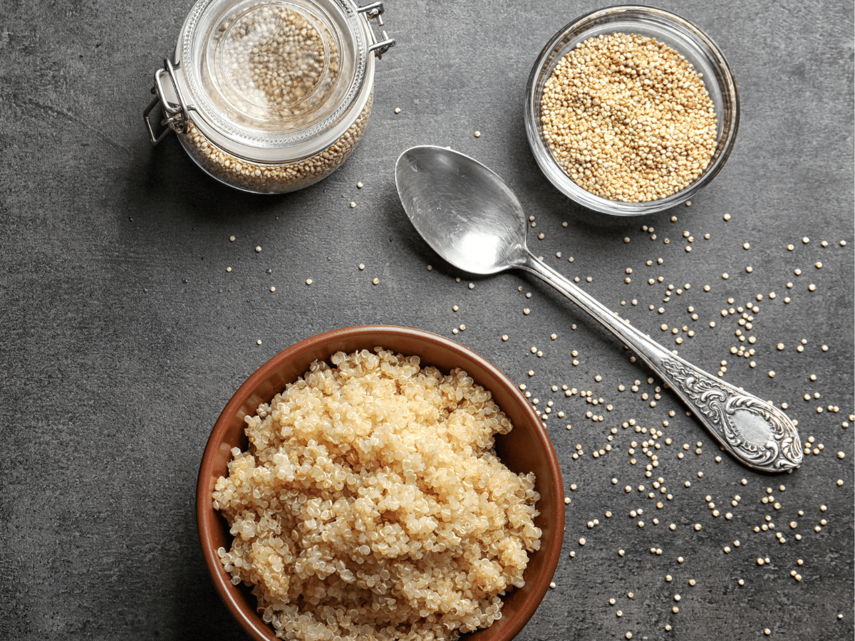 A bird's eye view of a bowl of cooked quinoa next to a spoon, jar, and bowl of uncooked quinoa against a grey background.
