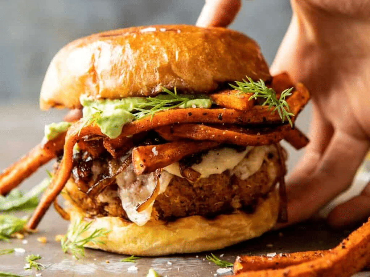 A close-up of a crispy quinoa burger topped with sweet potato fries, gruyere cheese, and caramelized onions.