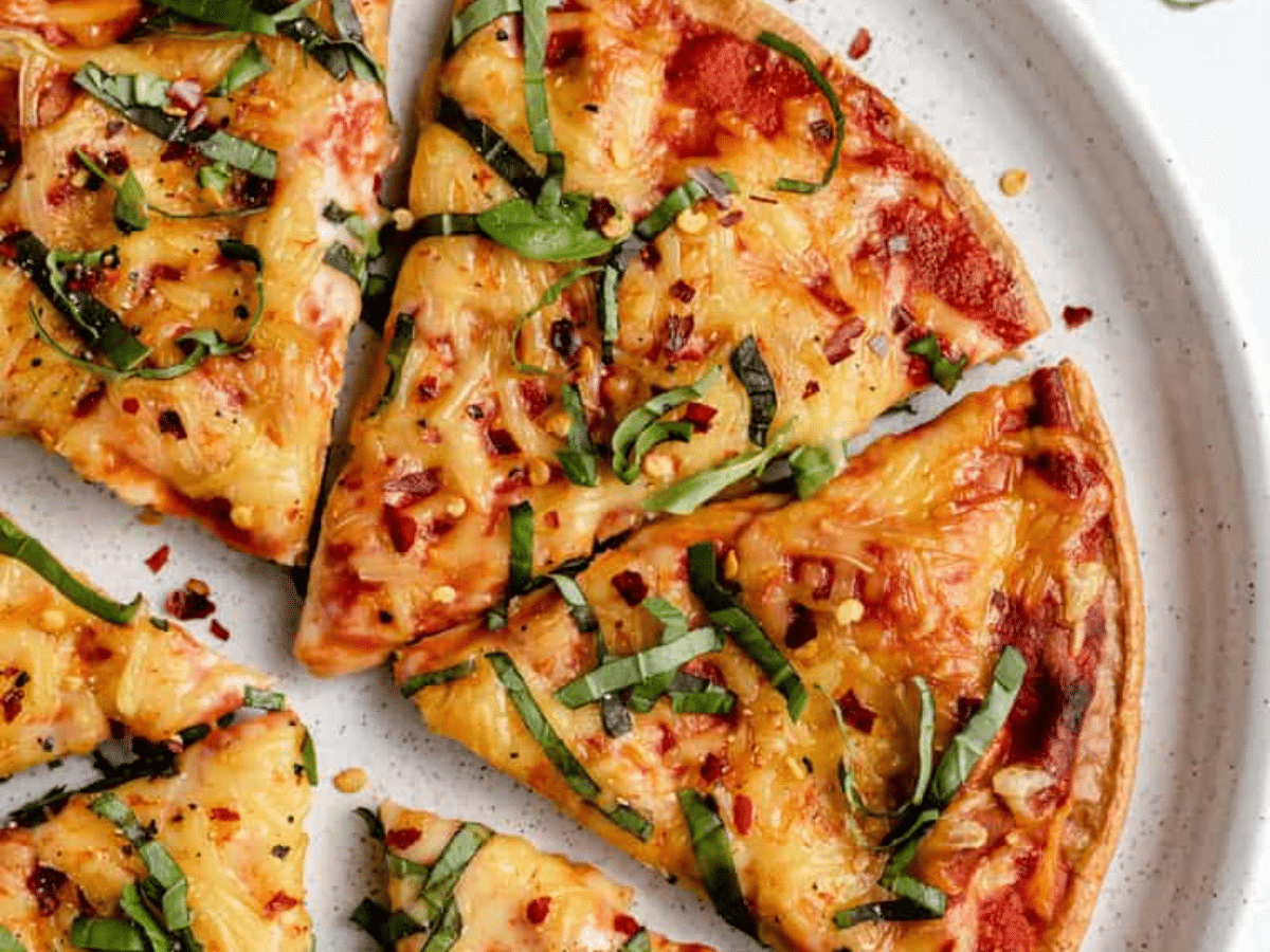 A close-up of cooked quinoa pizza crust  cut into slices and topped with cheese, crushed red pepper, and  fresh basil strips.