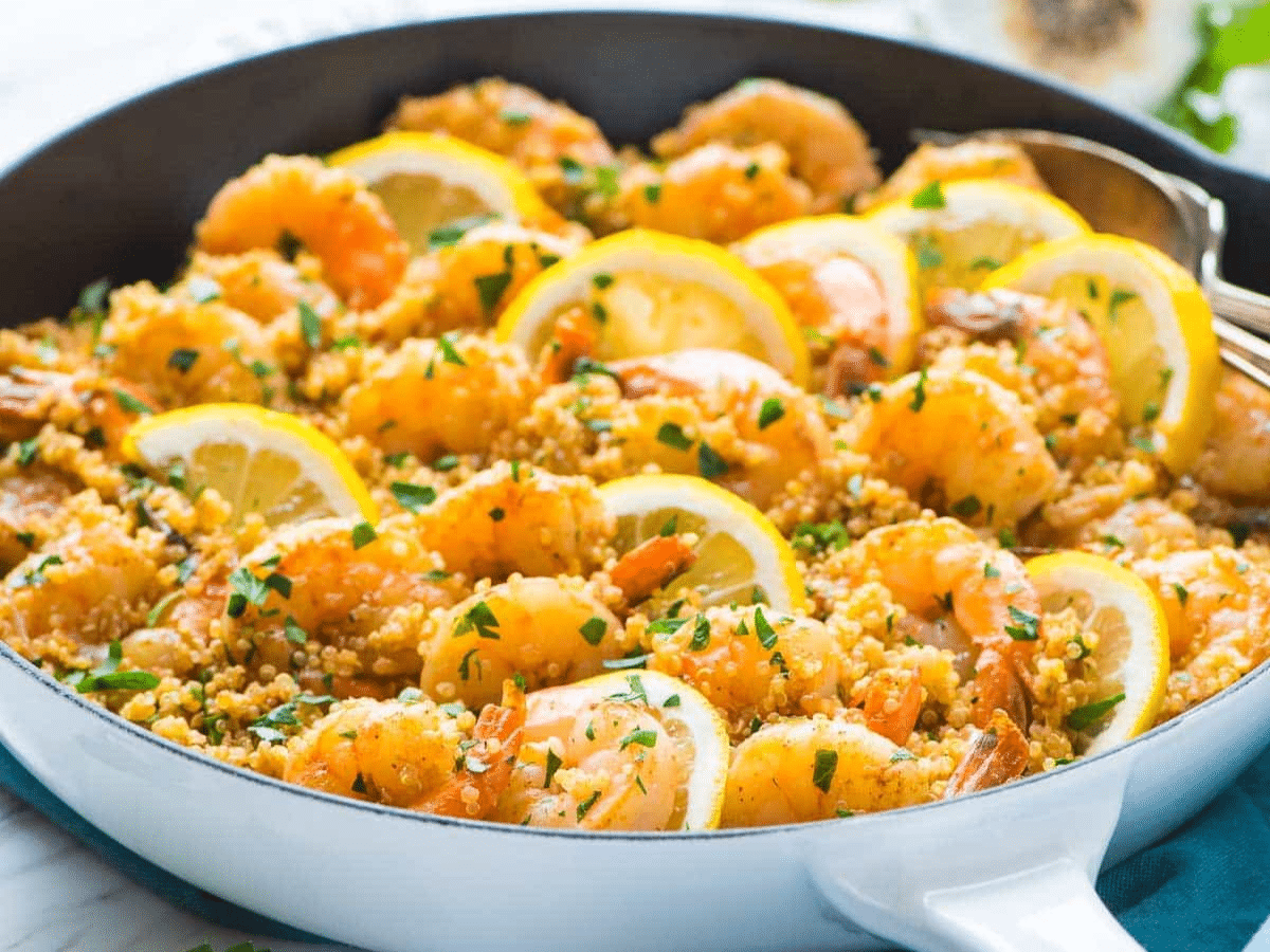 A close-up of a skillet filled with garlic shrimp, quinoa, herbs, and lemon slices.