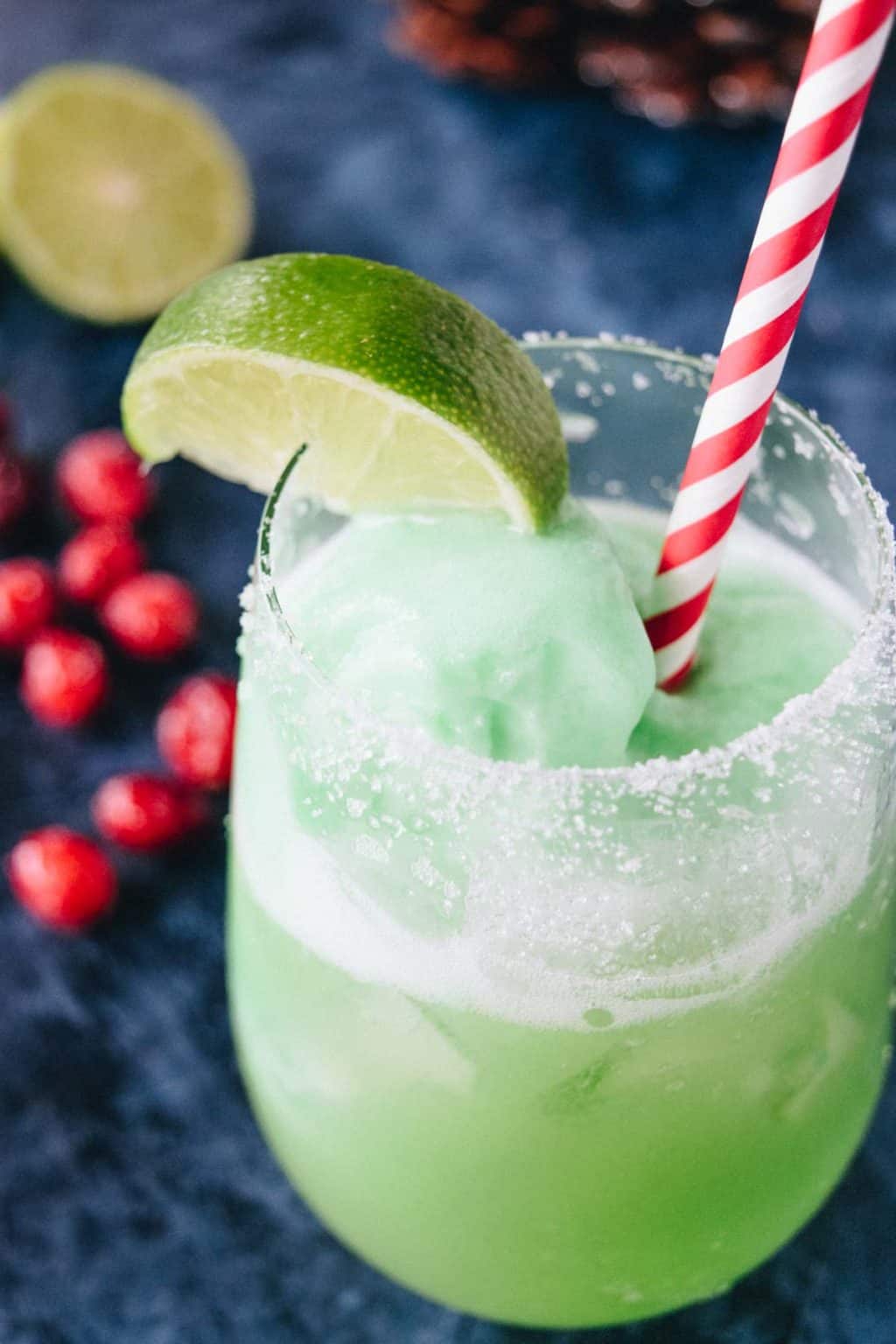 A clear glass filled with grinch punch and garnished with cranberries and a red and white candy cane striped straw