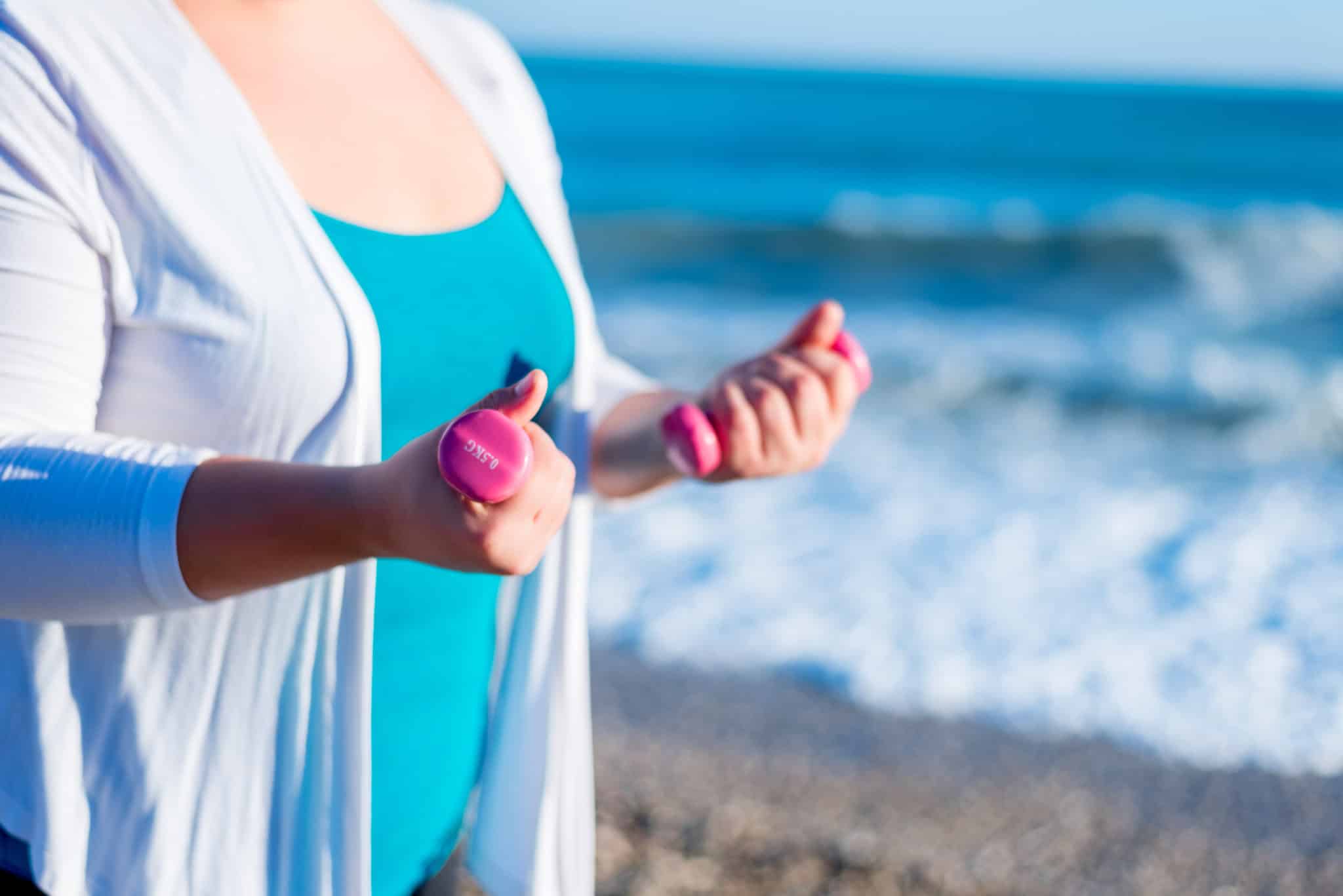 Woman in blue swimsuit and white shirt holding dumbbells on the beach.