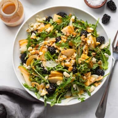arugula salad in a white bowl with pears, walnuts, gorgonzola, and blackberries.