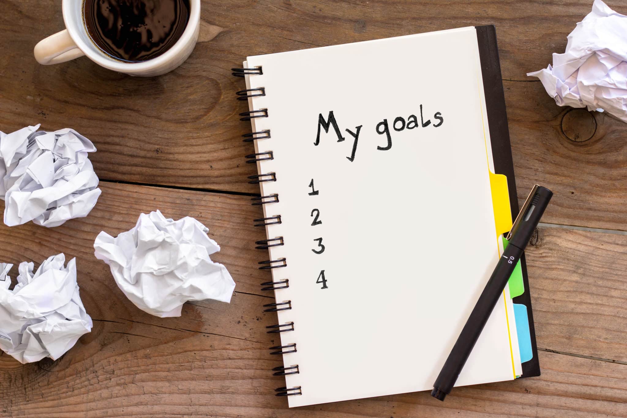 A notebook with "my goals" written across the top on wooden table with cup of coffee