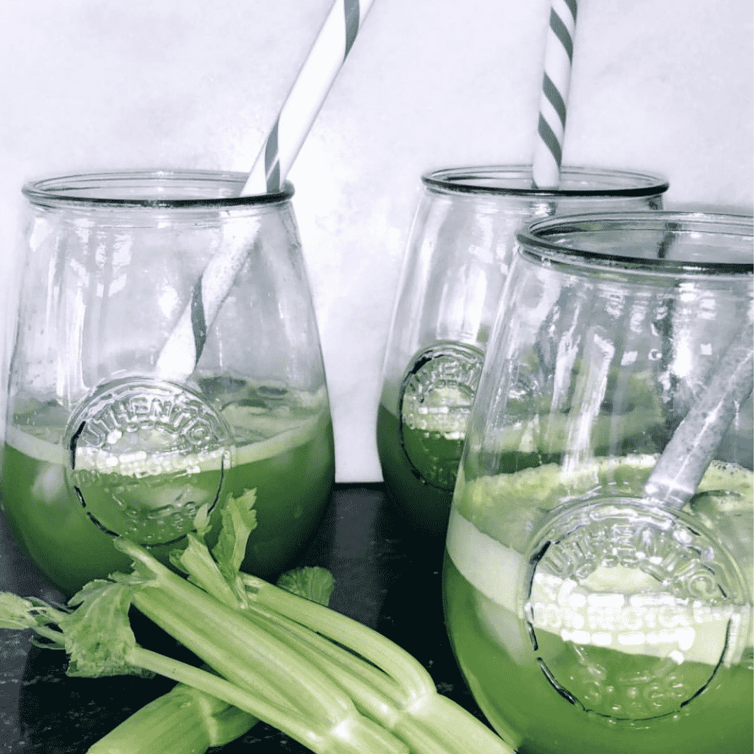 White background with three mason jar glasses filled with green juice. White and blue striped straws in each glass and a little stalk of celery lying on it's side.