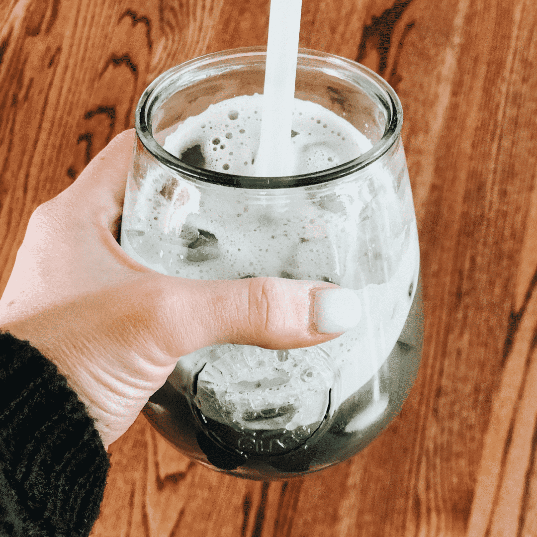 Women's hand holding a mason jar of green celery juice with a white straw over a dark wood floor.