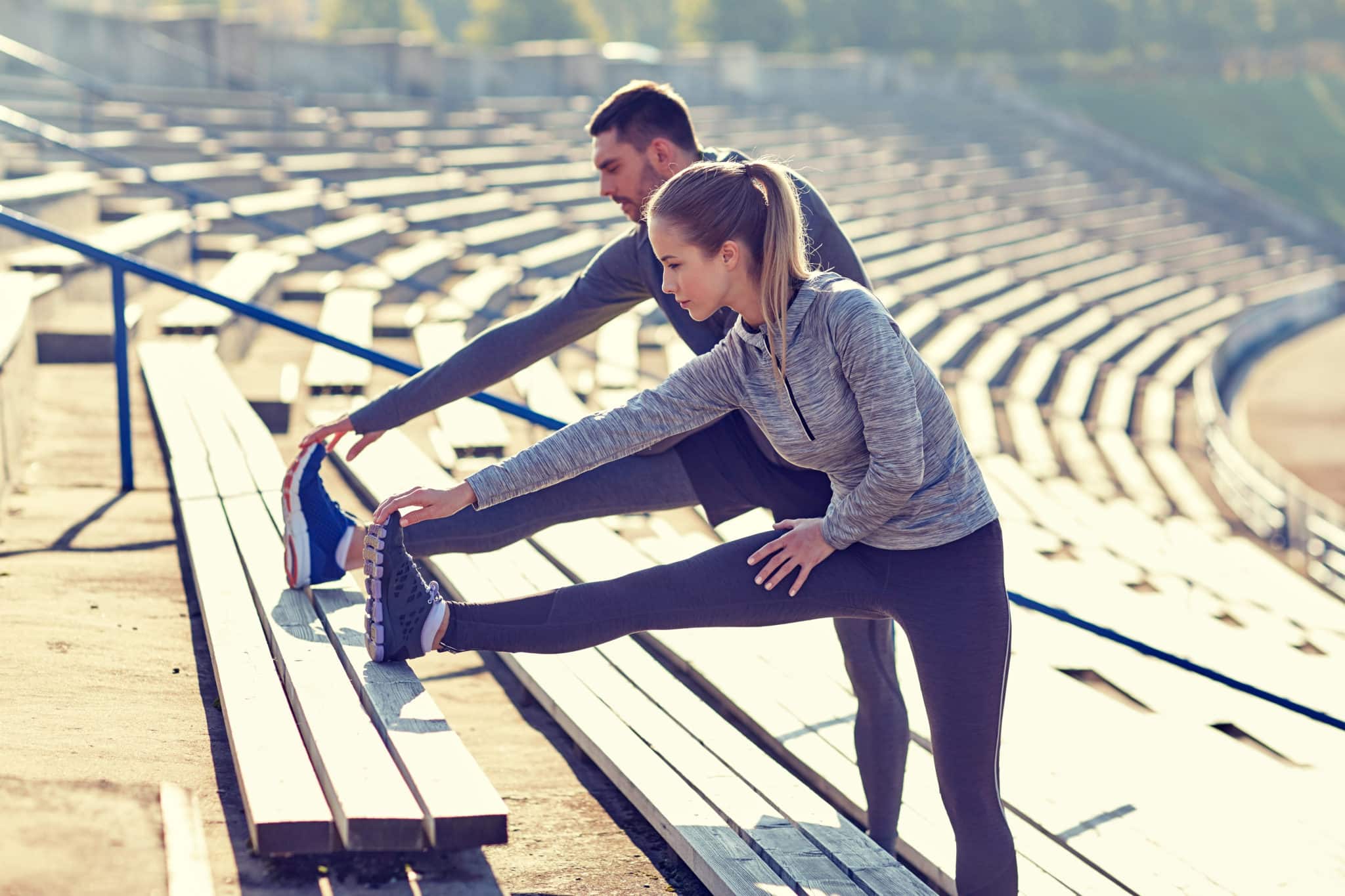 A man and a woman wearing athletic gear, stretching out their legs and reaching for their toes on bleachers.