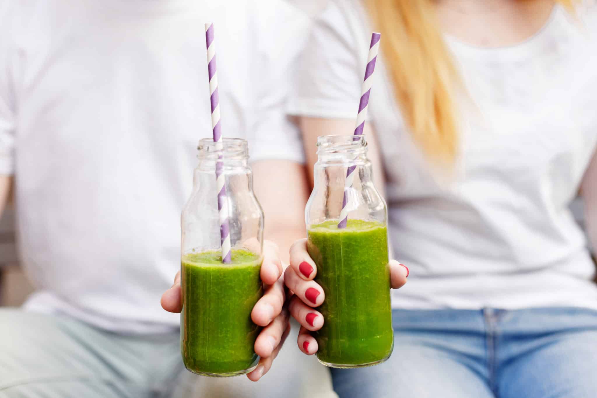 Two individuals in white t-shirts and jeans holding two clear bottles of green juice with white and pink striped straws.