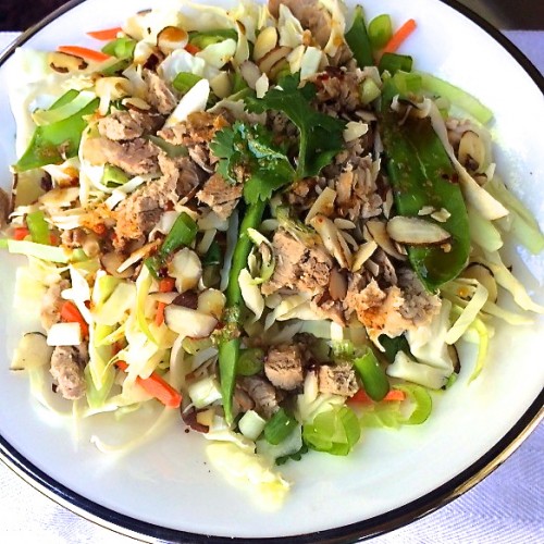 a white plate filled with a cabbage salad topped with sliced almonds, snow peas, carrots, green onions, and pulled pork