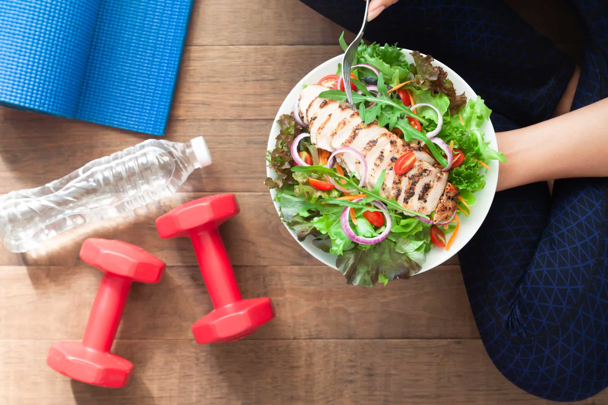 Woman holding bowl with green salad and chicken next to dumbbells, water bottle and yoga mat