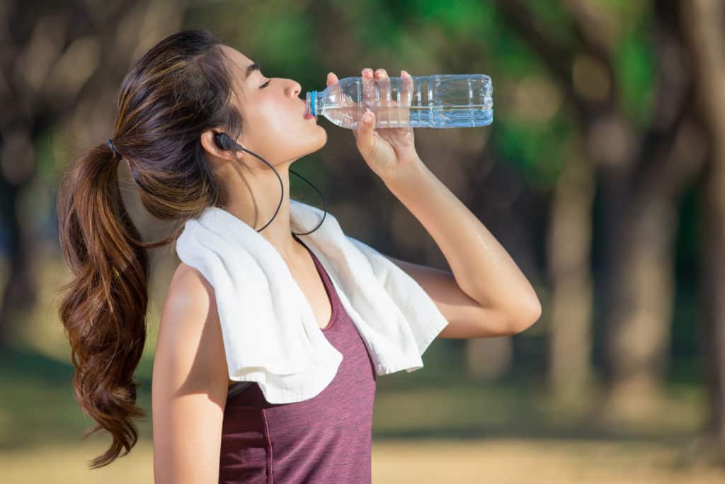 Young woman drinking bottle of water with towel around neck and earbuds in ears