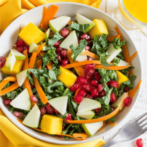 White bowl filled with kale salad including shaved carrots, pomegranate seeds, green apple and mango
