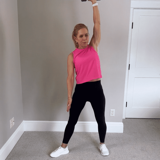Chris Freytag wearing a pink tank top and black leggings holding a 10 lb dumbbell above her head doing a dumbbell windmill. 