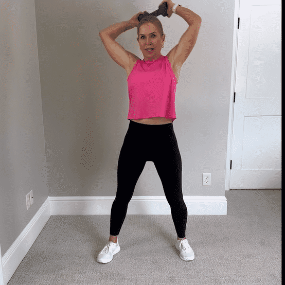 Chris Freytag wearing a pink tank top and black leggings performing a dumbbell halo using a dumbbell wrapping around your head. 