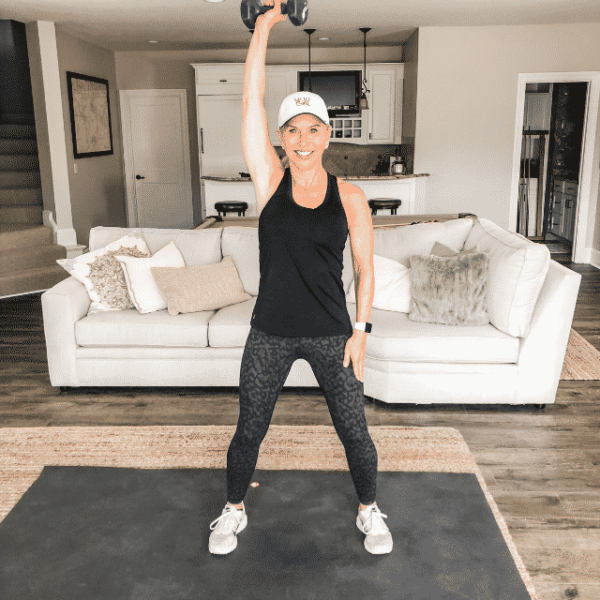 Chris Freytag wearing black and camo leggings doing a single arm overhead dumbbell press in basement.