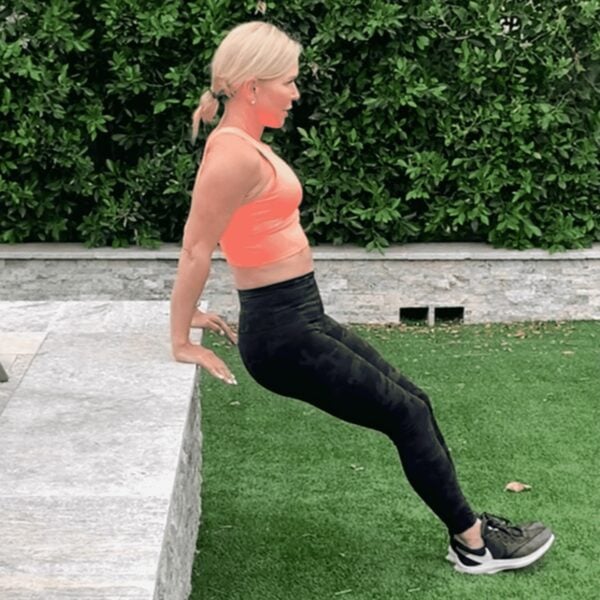 Chris Freytag outside wearing a bright orange tank top and black pants doing a tricep dip off of a marble ledge.