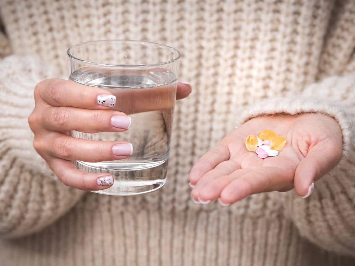 A woman with painted nails holding a glass of water and a handful of various vitamins.