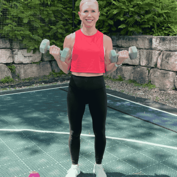 Chris Freytag wearing black leggings and orange top, holding an isometric hold of a bicep curl. Holding grey dumbbells in both hands.