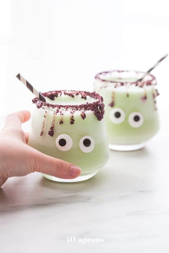 A green halloween "monsterita" drink recipe with googly eyes on a glass and dried blueberry crumbles around the rim