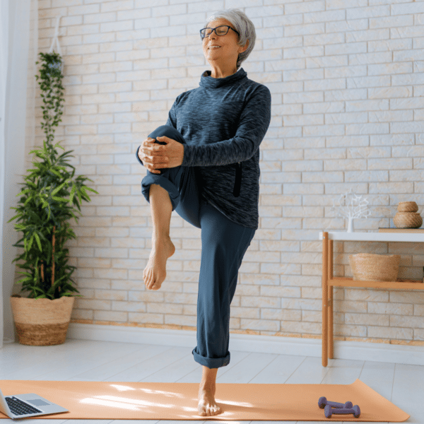 Senior woman performing hip strengthening exercises in living room with dumbbells and laptop