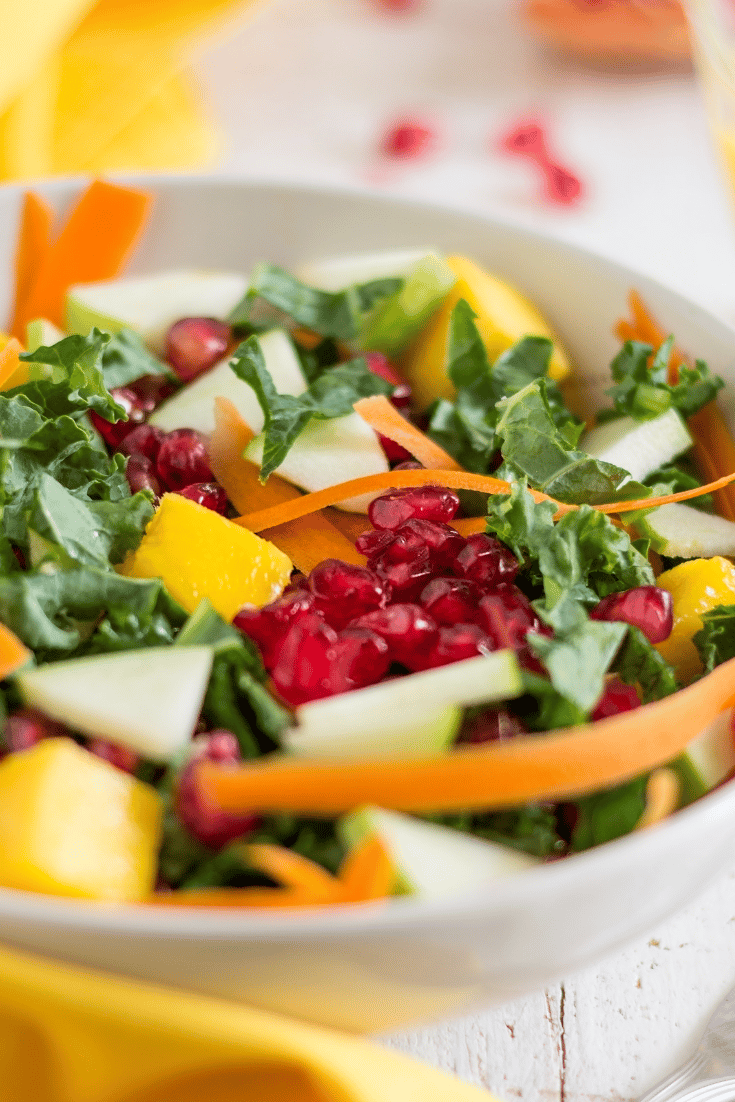 Crunchy kale salad in white bowl with shaved carrots, pomegranate seeds, mango and green apple
