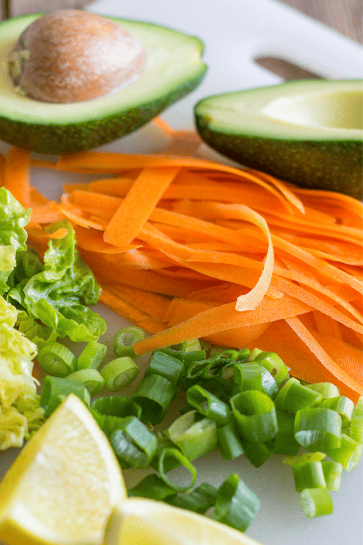Halved avocados, shaved carrots, wedges of lemon and green onions on cutting board