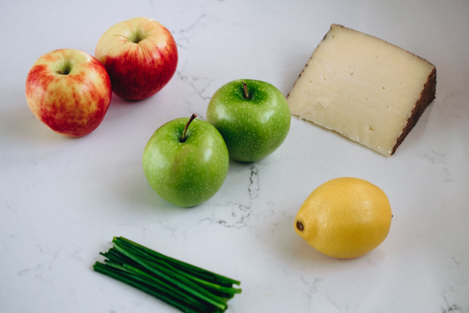 2 red apples, 2 green apples, block of manchego cheese, 1 lemon and chives on a white marble countertop
