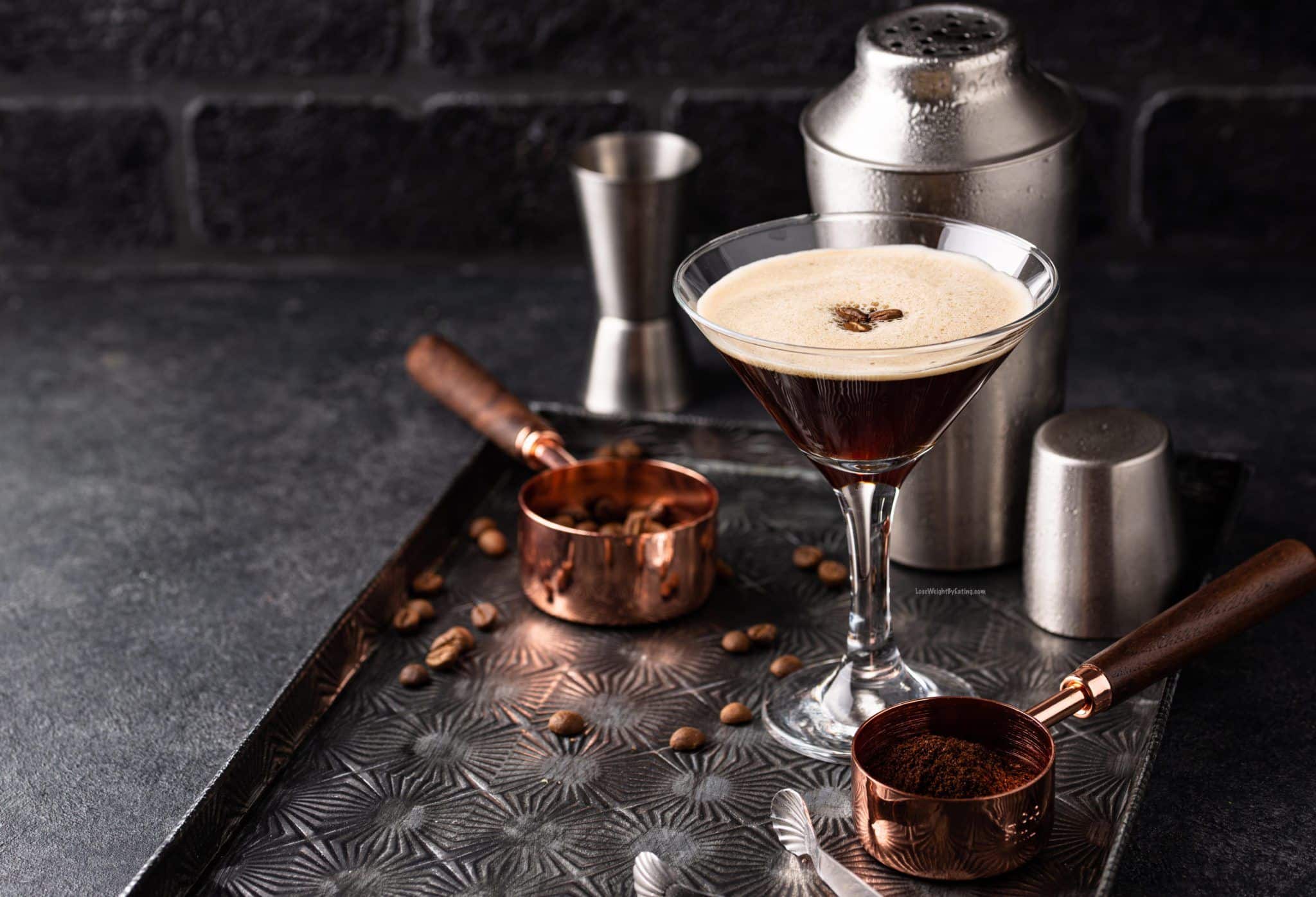 An espresso martini cocktail on a tray with measuring cups filled with ground and whole coffee beans and a cocktail shaker