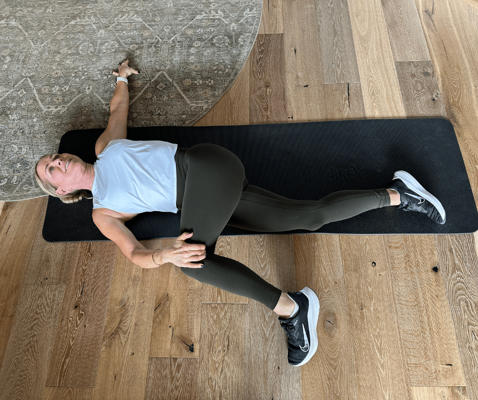 Chris Freytag wearing a white tank top and green leggings lying on a black yoga mat doing a lying spinal twist for sciatica relief.