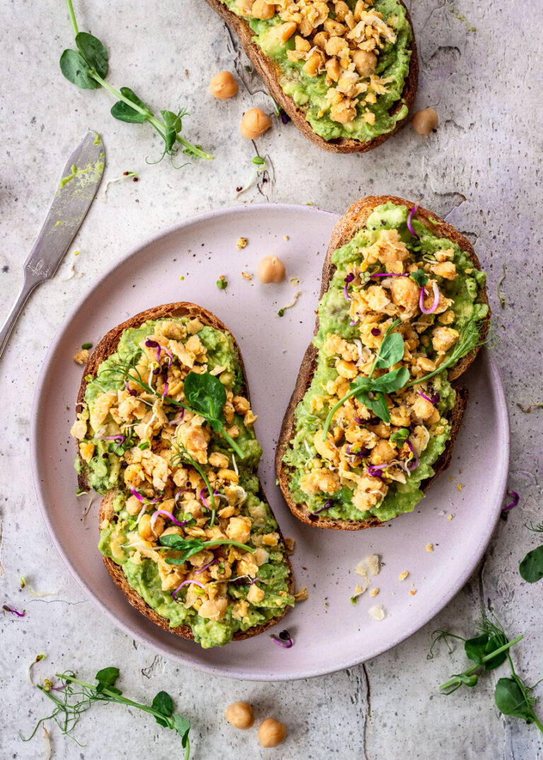 Two slices of avocado toast with smashed chickpeas on top on a neutral plate