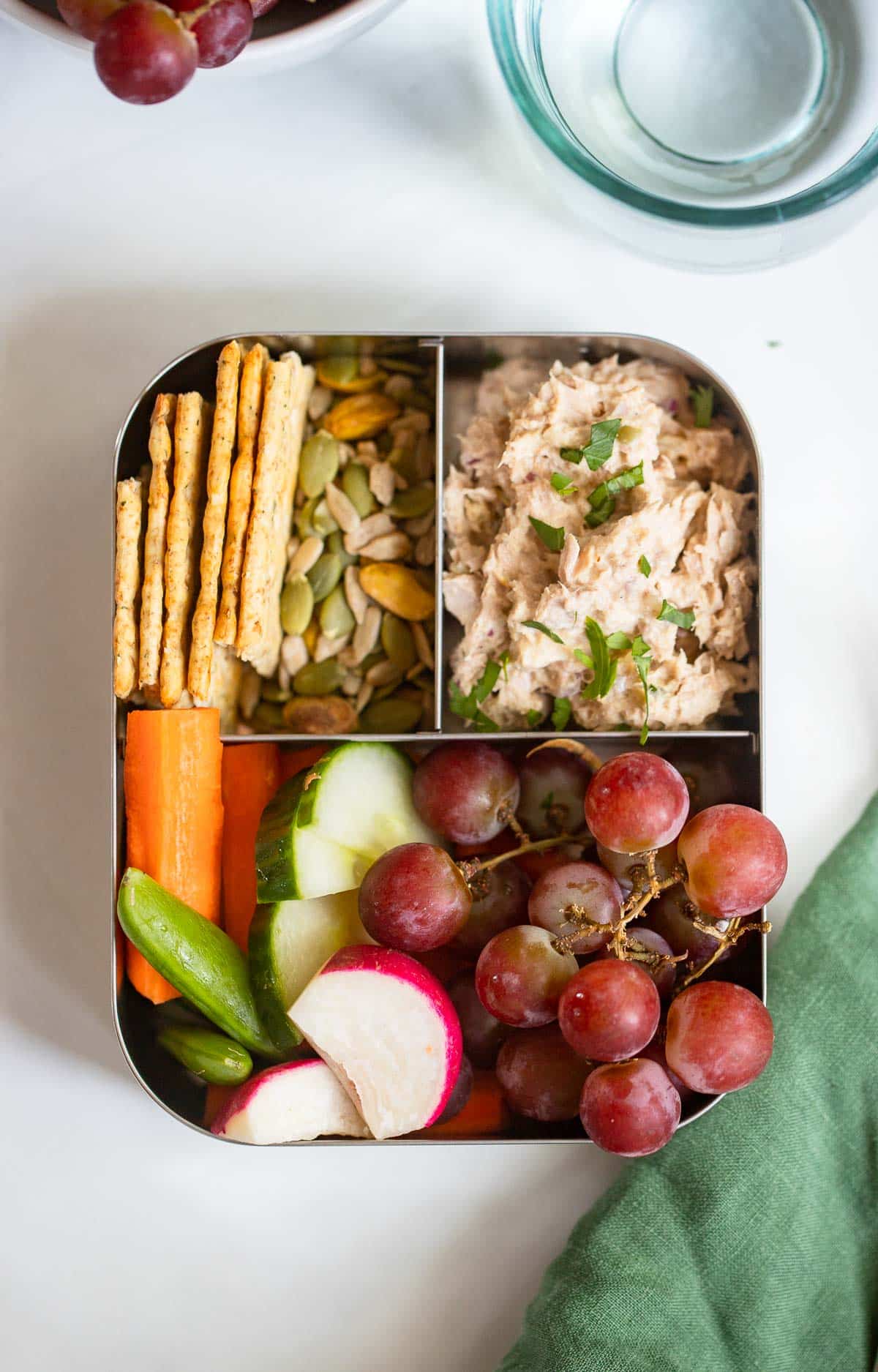 healthy tuna salad in a bento box with crackers, grapes, veggies, and trail mix.
