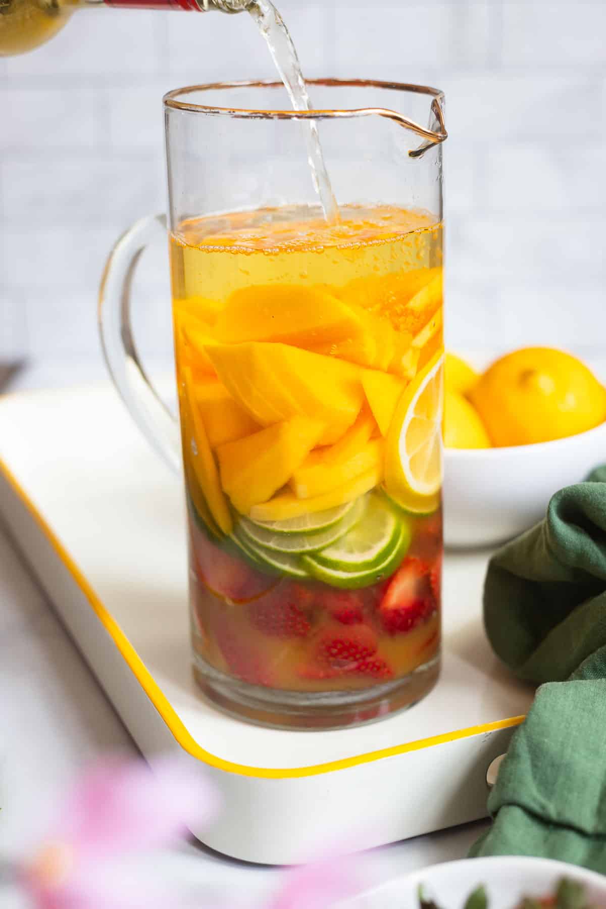 tall pitcher layered partially filled with sliced fresh fruit with a bottle of white wine being poured into it from the top.