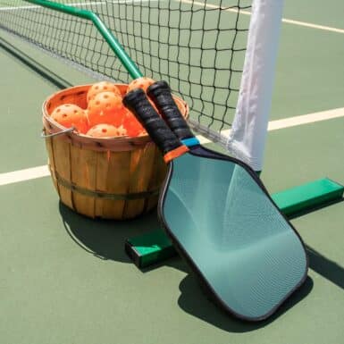 Two pickleball paddles and a basket of pickleballs on a pickleball court next to the pickleball net.