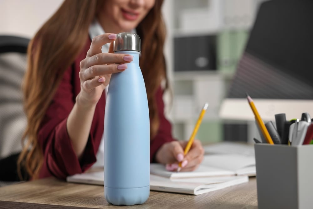 Woman at a desk writing in a notebook while holding a large blue thermos in the other hand.